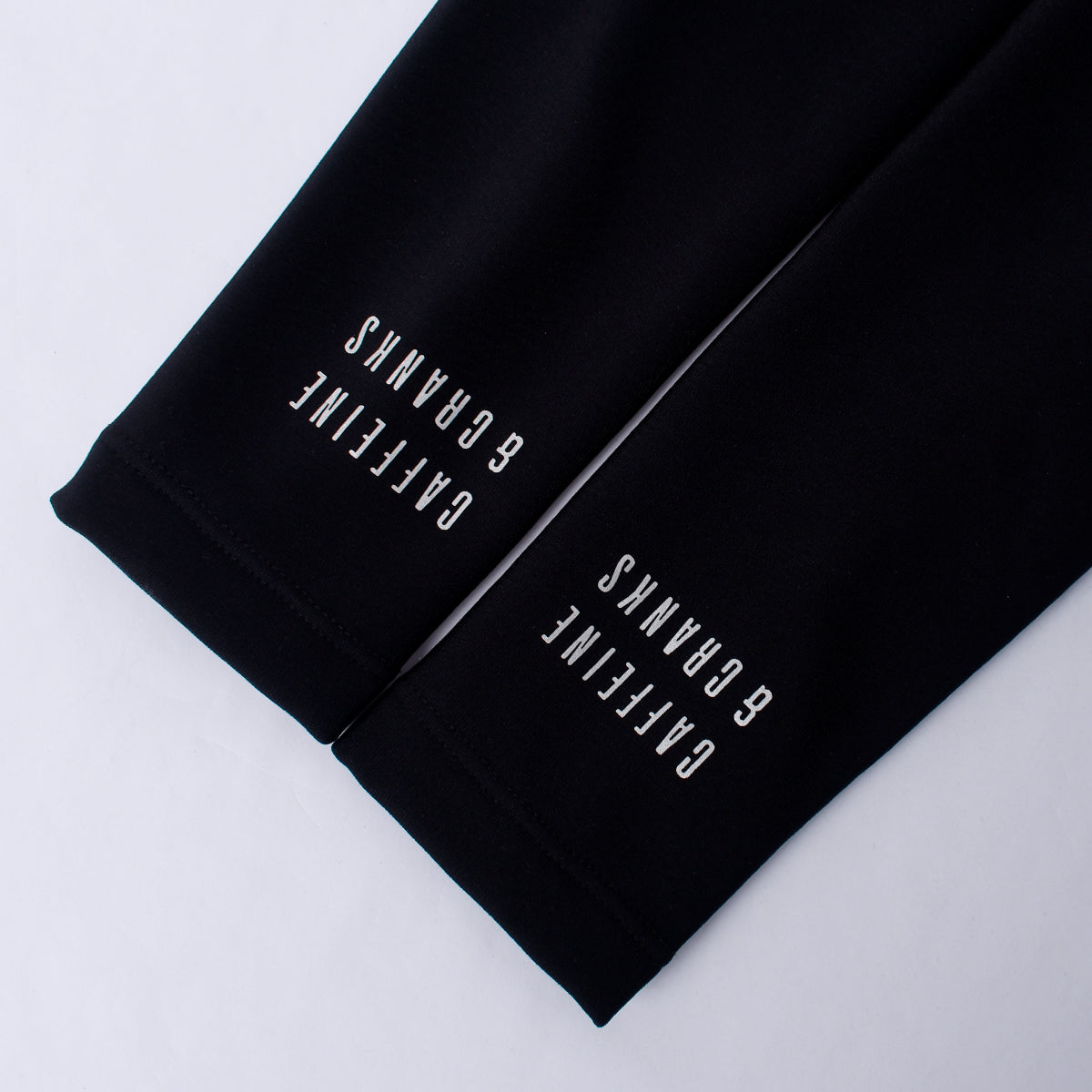 Close-up view of black thermal arm warmers with white "CAFFEINE & CRANKS" text.