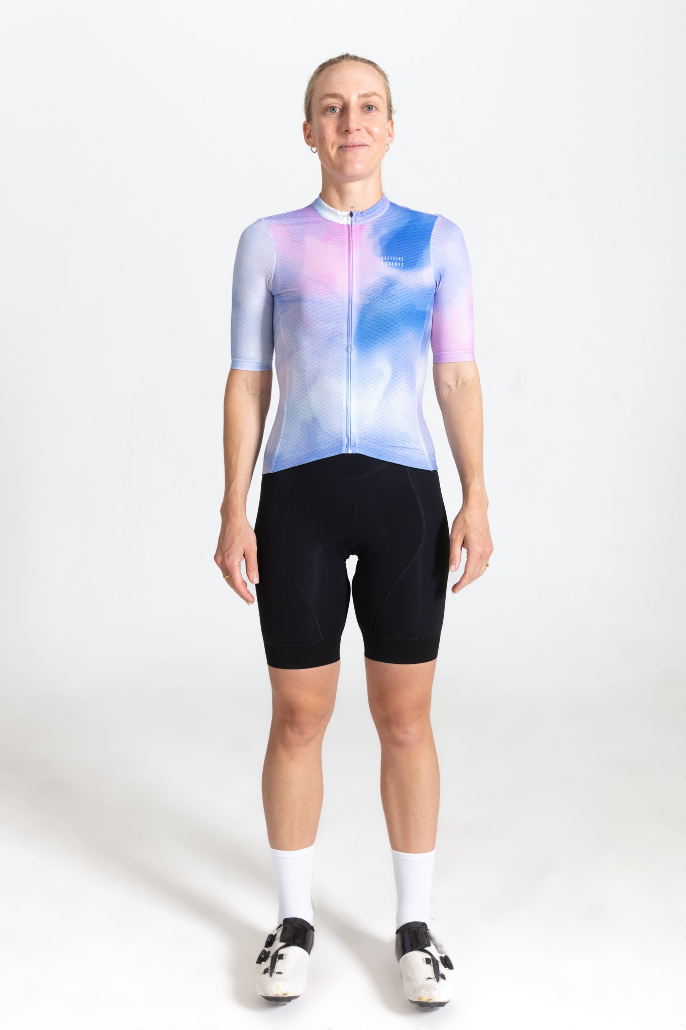 Woman in C&C Coral Jersey with pink & blue cloud pattern, short sleeves, black cycling shorts, white socks, and cycling shoes.