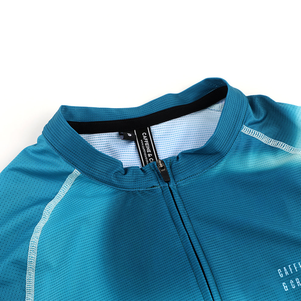 Close-up of C&C Flux Jersey collar on men's cycling shirt. Features teal gradient & white stitching.