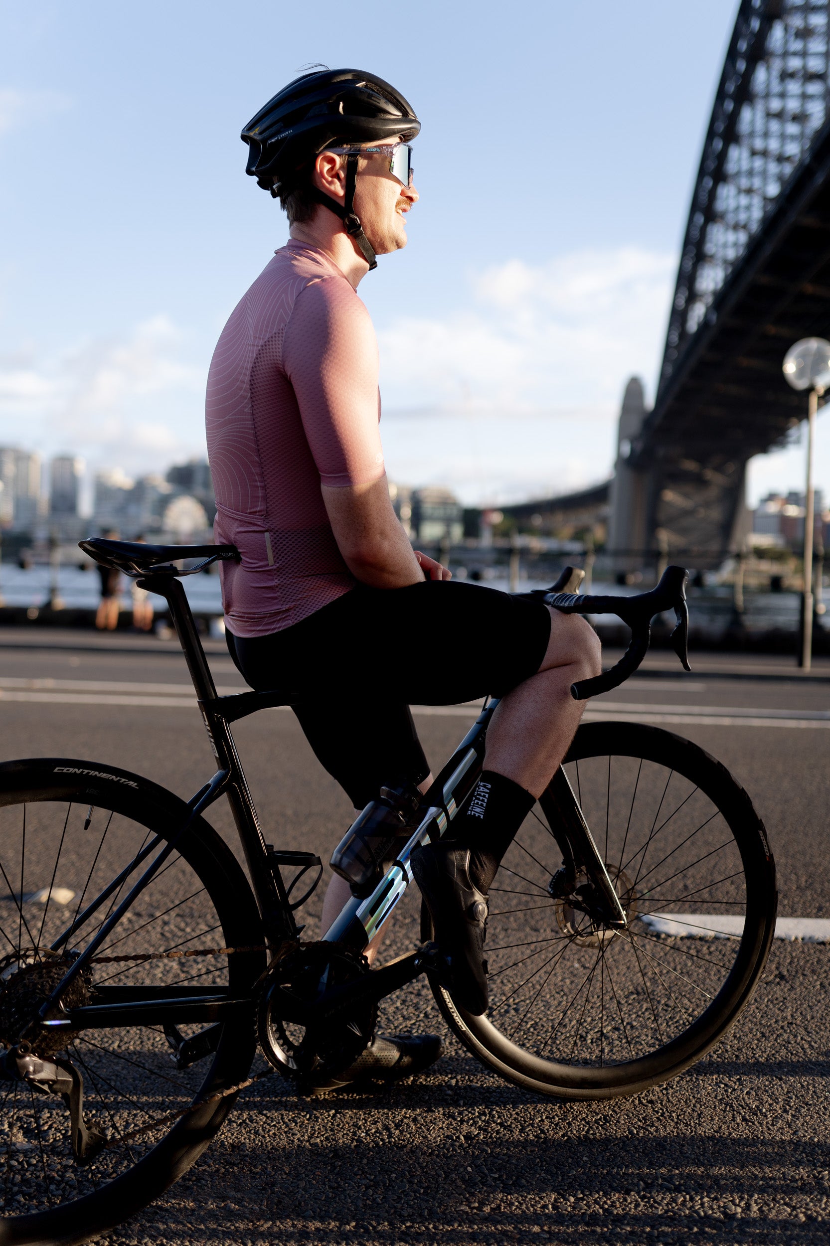 Man in C&C Garnet Topo Jersey and black shorts on a bike, with sunset and city skyline.