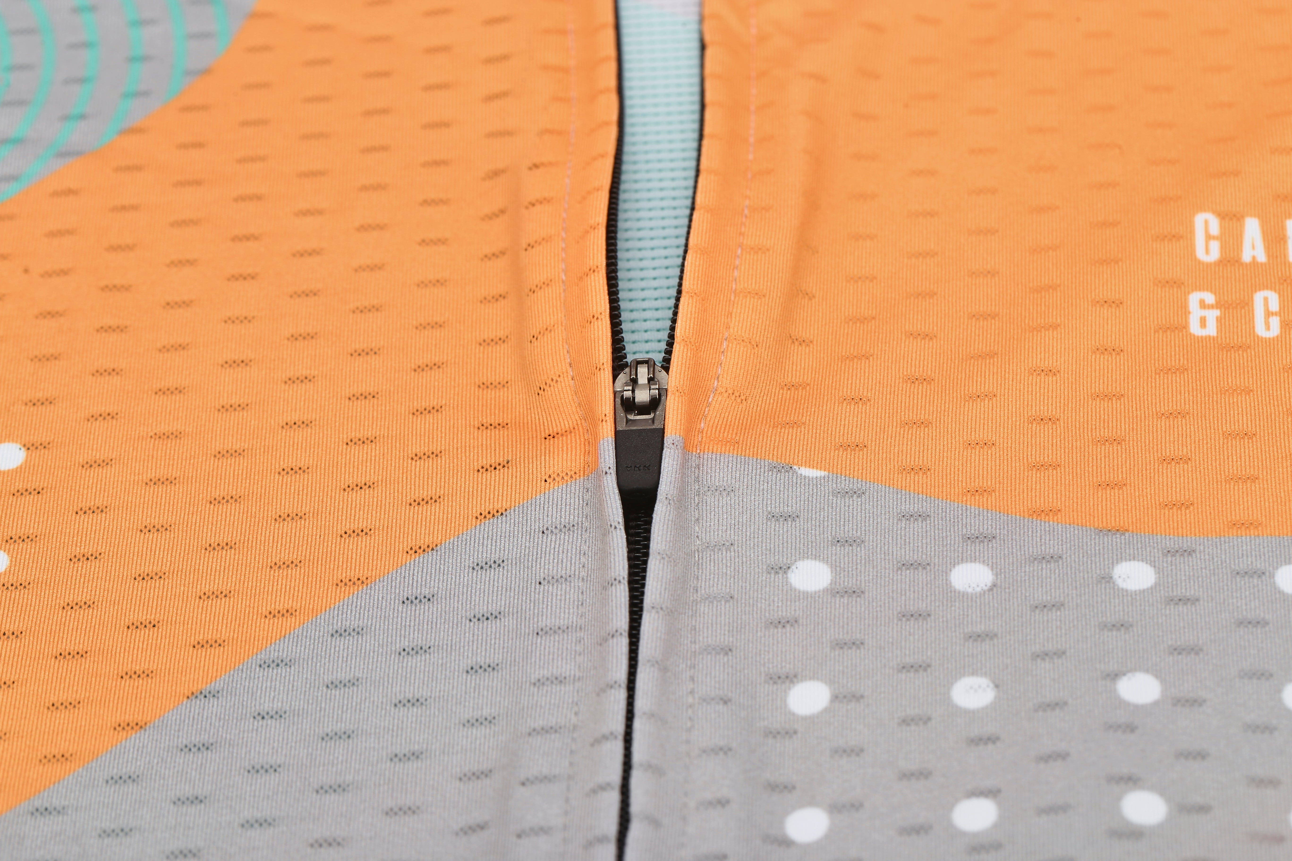 Close-up of C&C Memphis Light Jersey, showing orange fabric with grey & teal accents and a zipper.
