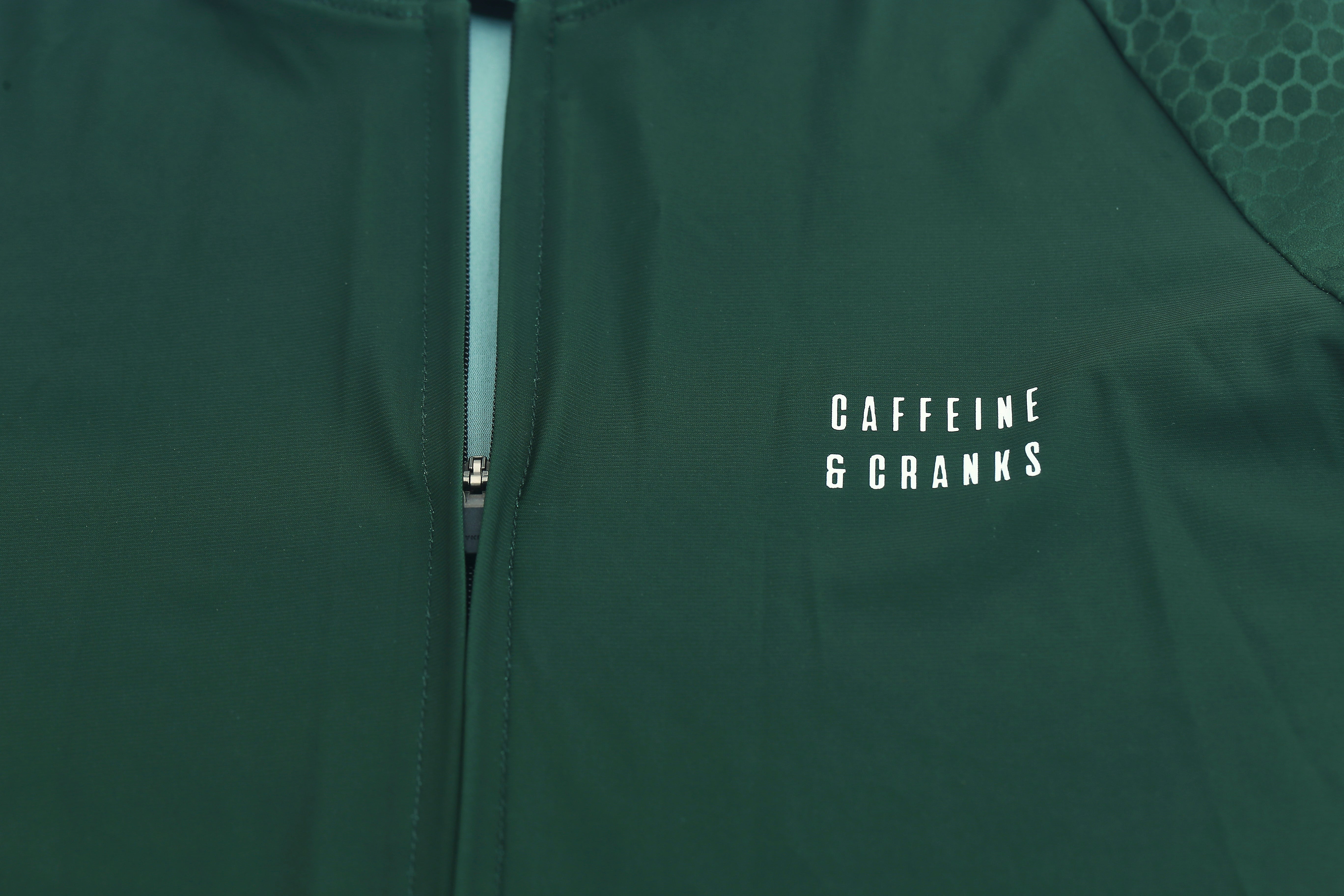 Close-up of forest green women's Pinnacle jersey with white "CAFFEINE & CRANKS" logo and honeycomb detail.